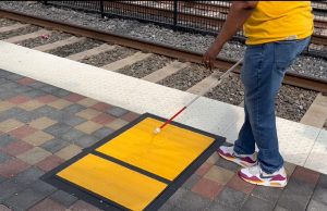 A person uses a white cane to locate a yellow tactile path beside a train platform edge.