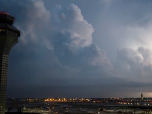 An airport control tower stands beside an airfield under large storm clouds, with aircraft and terminal areas visible below.