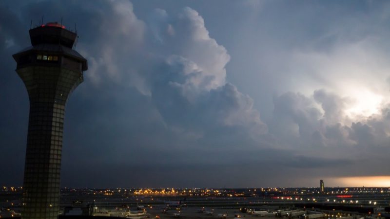 An airport control tower stands beside an airfield under large storm clouds, with aircraft and terminal areas visible below.