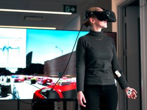 A woman wears virtual reality goggles with a cable connected to equipment outside the frame and has a galvanic skin response sensor attached to her lower arm and a sensor on her palm. Her hand holds a small device linked to the sensor. Behind her is a large screen that shows a simulated two-way city street with many cars and buildings. The woman faces slightly to the side and appears to engage with the virtual scene.
