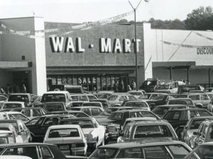 A crowded parking lot filled with 1980s cars stands in front of a Wal-Mart store with Discount City signage.