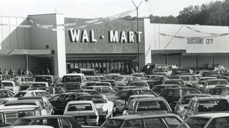 A crowded parking lot filled with 1980s cars stands in front of a Wal-Mart store with Discount City signage.