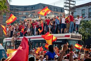 A group of World Cup soccer fans stand on top of a bus and wave Spanish flags. More people stand around the bus and hold flags in a crowded street.