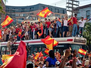 A group of World Cup soccer fans stand on top of a bus and wave Spanish flags. More people stand around the bus and hold flags in a crowded street.