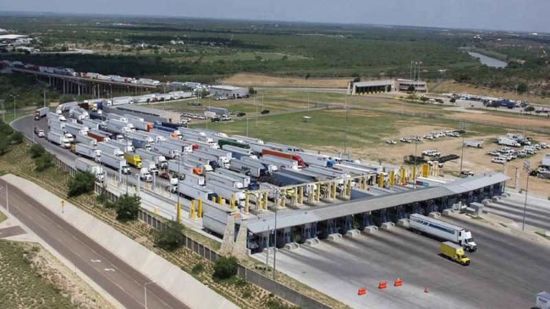 The aerial view of the World Trade International Bridge shows many trucks lined up in multiple lanes. The trucks wait at a series of inspection booths with canopies and equipment. The lanes extend toward the bridge structure in the distance. A few trucks move through open lanes while many others remain stopped. The area around the bridge includes paved roads, open land, parking areas, and scattered buildings. Trees and a river appear in the background along with wide stretches of land.